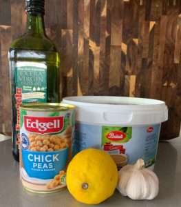 An image of the ingredients for hummus including a tin of chickpeas, a lemon, a bulb of garlic, a bottle of olive oil against a wooden background,
