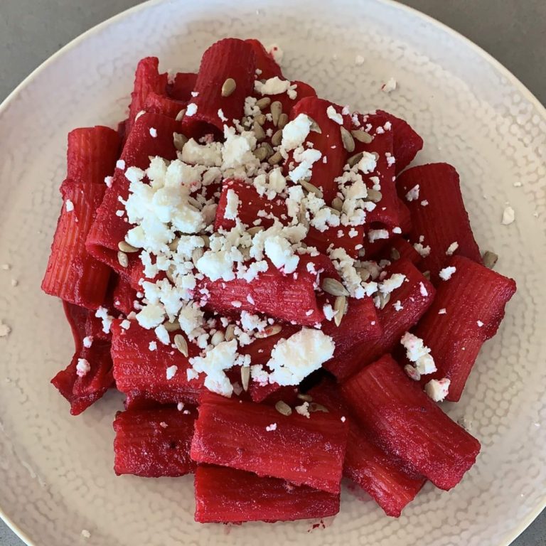 A plate of rigatoni pasta coated in a bright pink beetroot sauce topped with sunflower seeds and feta.