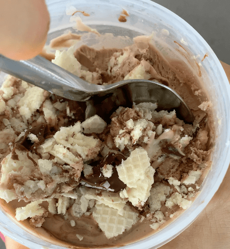 A tub of Nutella Ice Cream being scooped into topped with chopped wafers and Nutella