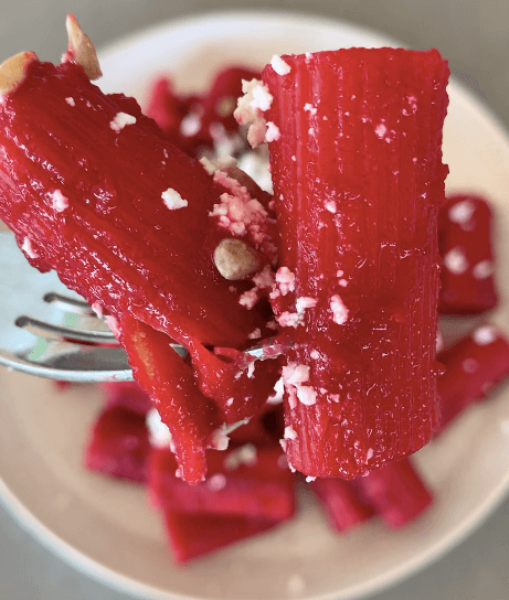 A close up of a forkful of rigatoni pasta coated in a bright pink beetroot sauce topped with sunflower seeds and feta.