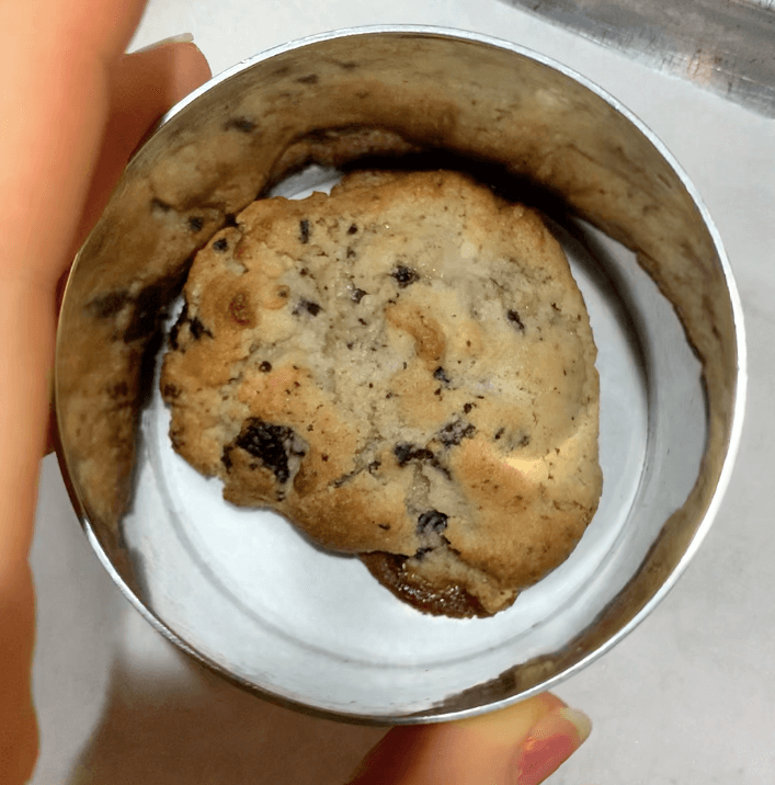 An unevenly shaped chocolate chip cookie on a baking tray with a large cookie cutter over the top of it