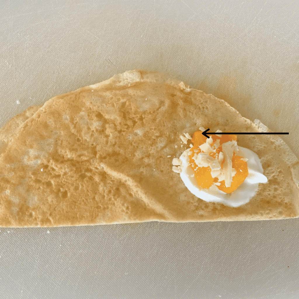Half a crepe on a chopping board with yoghurt, diced mango and chopped white chocolate