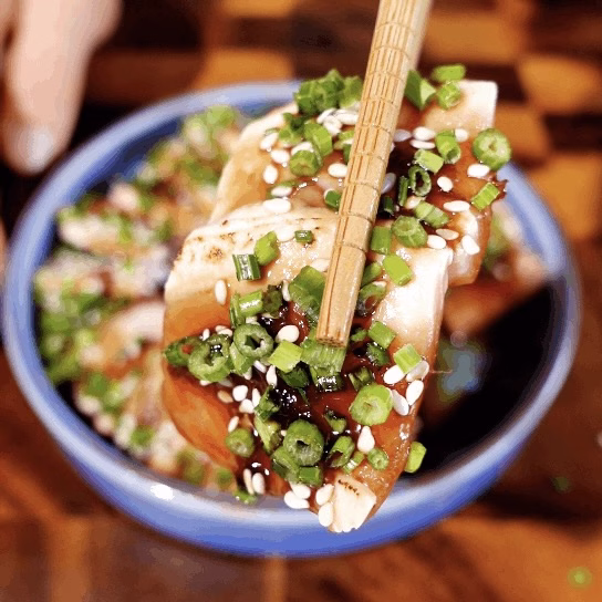A close-up shot of wooden chopsticks lifting a single slice of torched salmon covered in sticky glaze, toasted sesame seeds, and green spring onions.